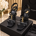 A pair of black Saint Laurent high-heeled sandals with gold-tone YSL logo hardware, displayed on top of black Saint Laurent boxes with a branded shopping bag and a vase of white flowers in the background.