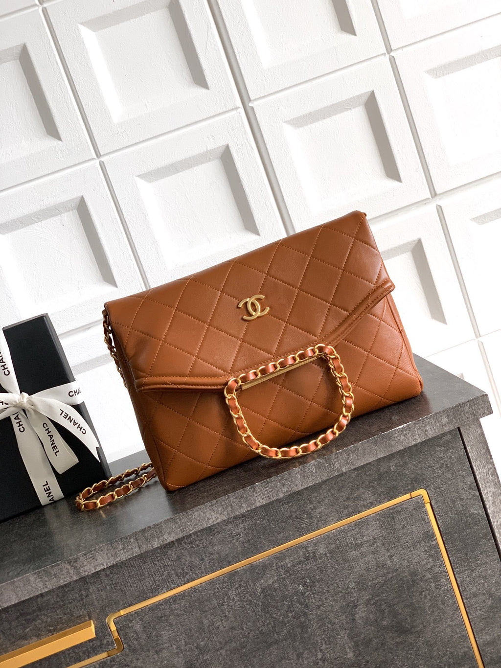 Brown quilted handbag with gold chain on a gray surface with a white geometric patterned wall in the background.