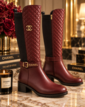 A pair of Chanel knee-high boots in deep burgundy with black side panels, quilted stitching on the shaft, a gold-tone CC logo, and a matching gold buckle strap, displayed on a marble surface with Chanel-branded books and a candle.