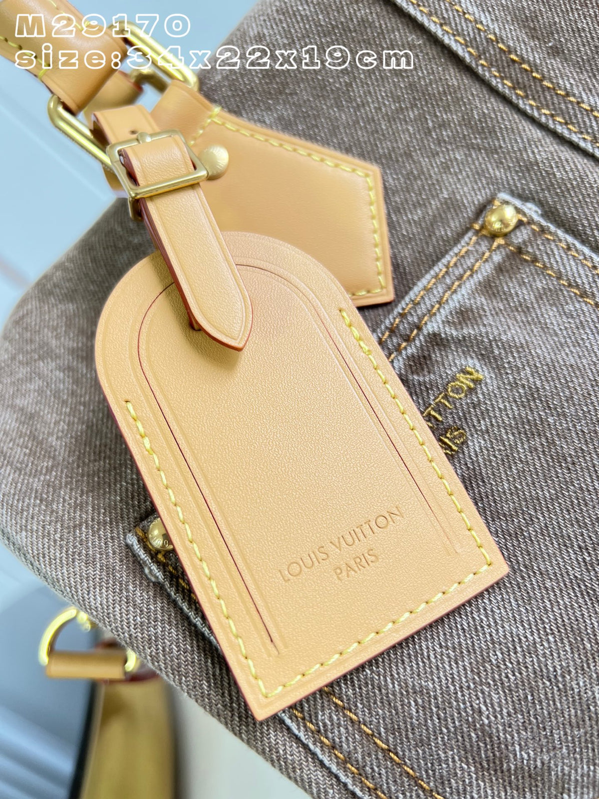 A close-up of a Louis Vuitton leather luggage tag attached to a gray denim bag. The tag is a light tan/beige color with gold-tone hardware and yellow stitching, and the Louis Vuitton name is embossed on the bottom right.