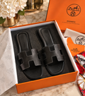 A pair of black leather Hermes slide sandals with signature H-ear straps, presented in an orange Hermes box with branded tissue paper, dust bag, and orange box on a marble surface.