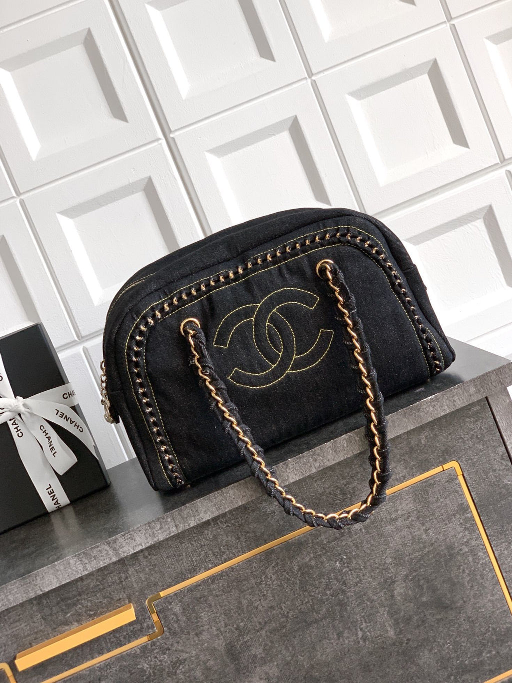 A black denim shoulder bag with gold-tone hardware, featuring a prominent interlocking CC logo on the front and a braided chain handle, displayed on a dark gray cabinet against a white tiled wall.
