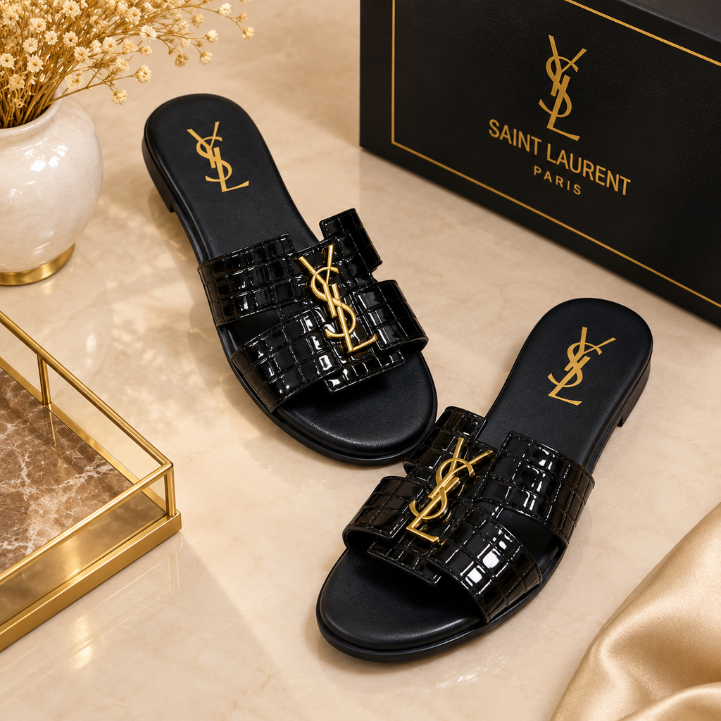 A pair of black Saint Laurent slide sandals with gold-tone YSL logo hardware, displayed on a beige marble surface next to their black branded box.