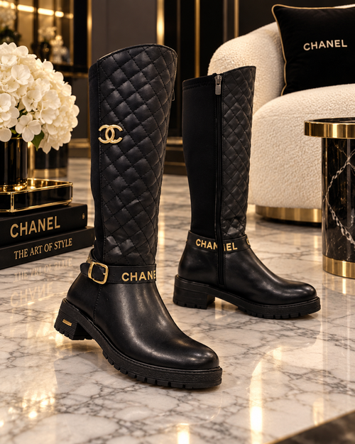 A pair of black Chanel knee-high boots with quilted panels, gold-tone hardware, and the Chanel logo and wordmark, displayed on a marble surface in a luxury interior setting.