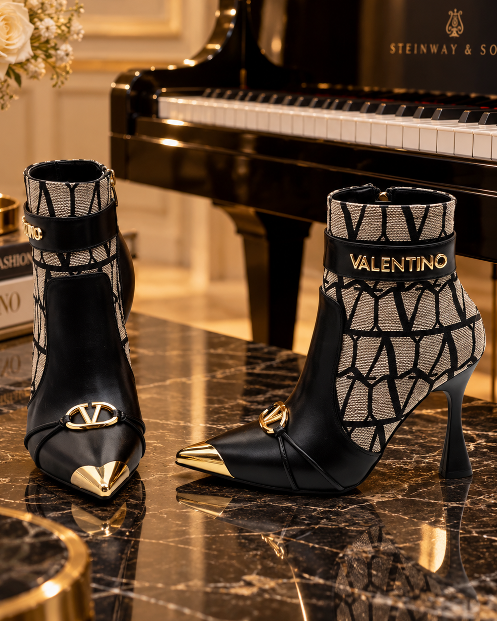 A pair of Valentino pointed-toe ankle boots with black leather and a beige geometric pattern, displayed on a polished black marble table next to a black Steinway & Sons grand piano.