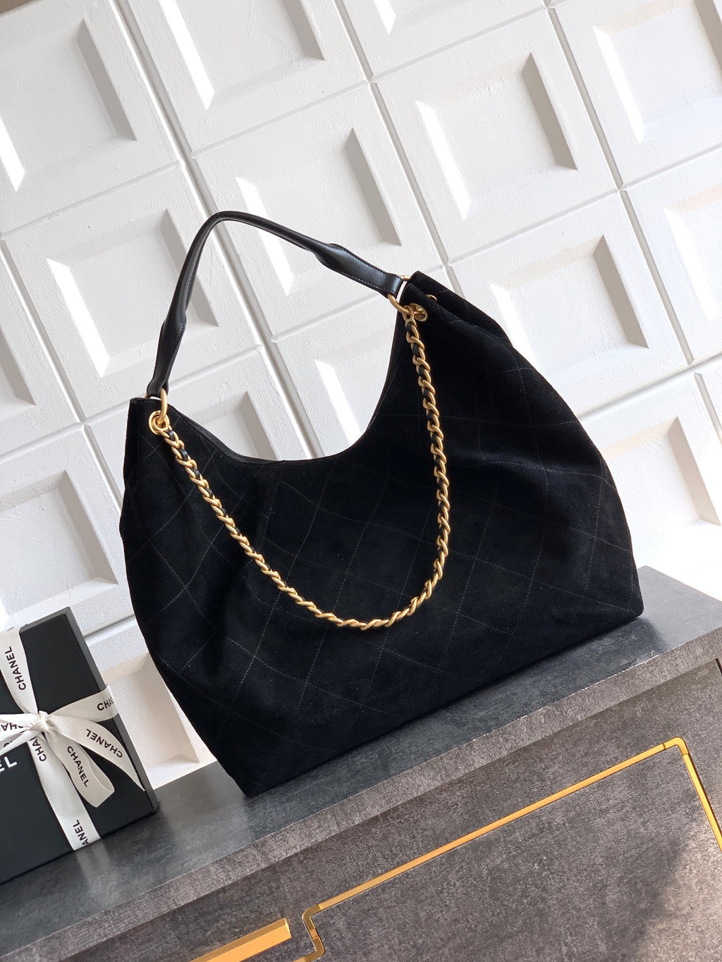 A black quilted suede hobo handbag with a gold chain strap, resting on a gray stone ledge next to a black Chanel gift box against a white tiled wall.
