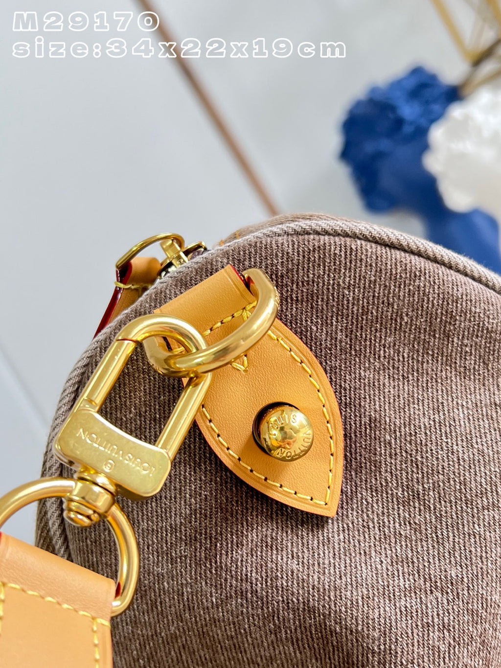 Close-up of a brown woven fabric bag with a tan leather strap tab and gold-tone hardware, including a D-ring clasp engraved with 'Nagasaki' and a round snap button.