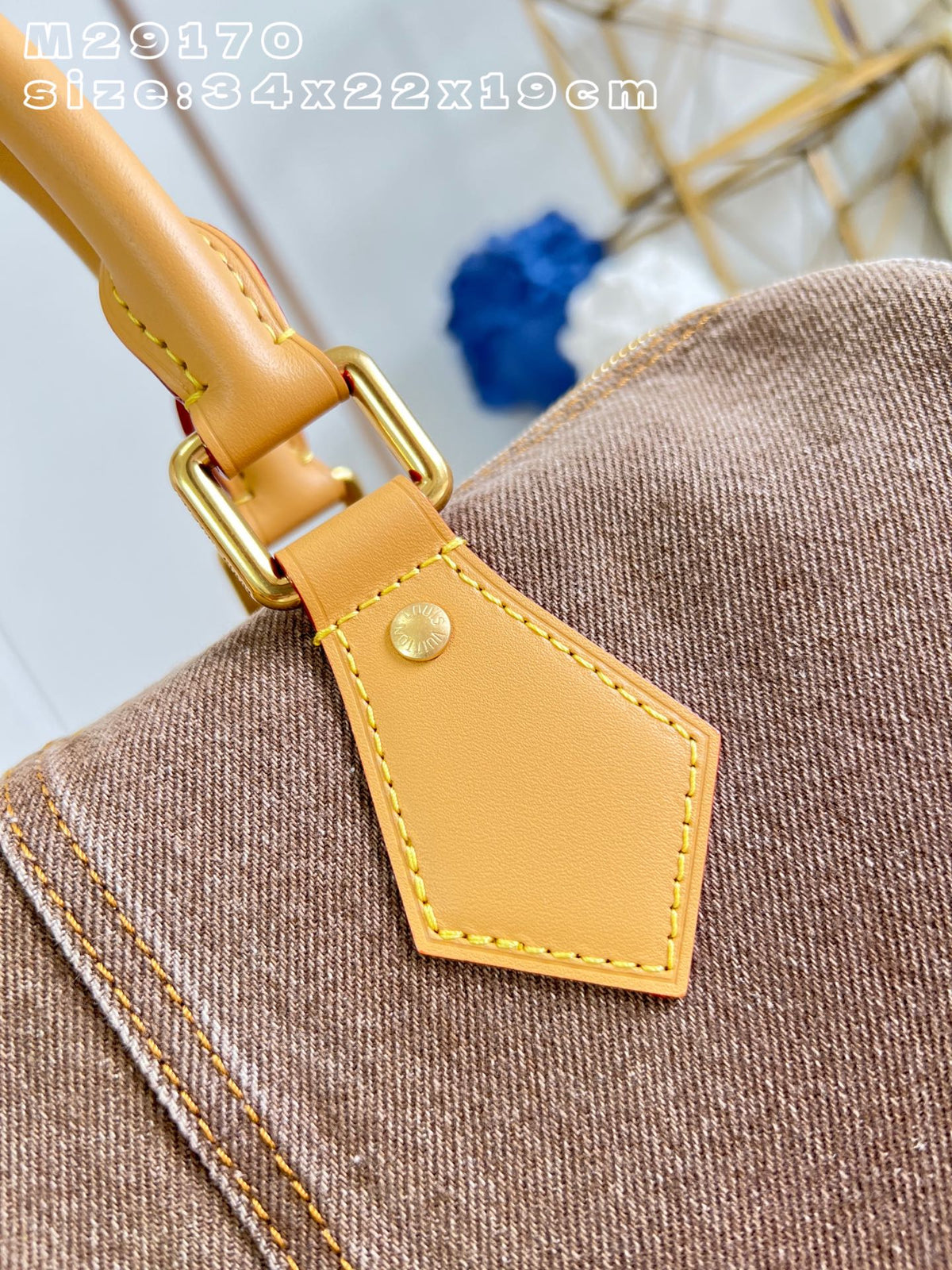 Close-up detail of a brown canvas handbag with a tan leather strap tab and gold-tone hardware, showing the texture of both materials. White text overlay in the top left reads 'M29170 size:34x22x19cm'.