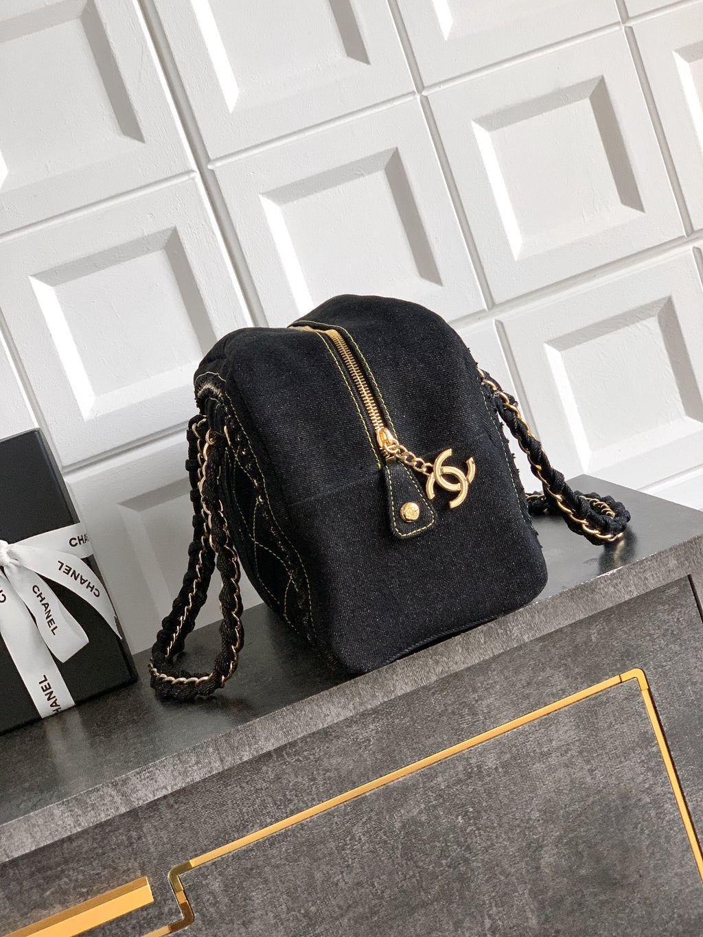 A black Chanel mini backpack with gold hardware and a braided chain strap, resting on a dark gray surface in front of a white tiled wall.