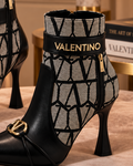 A pair of Valentino pointed-toe ankle boots with black leather and a gray geometric pattern, displayed on a pink velvet ottoman in an elegant room setting.