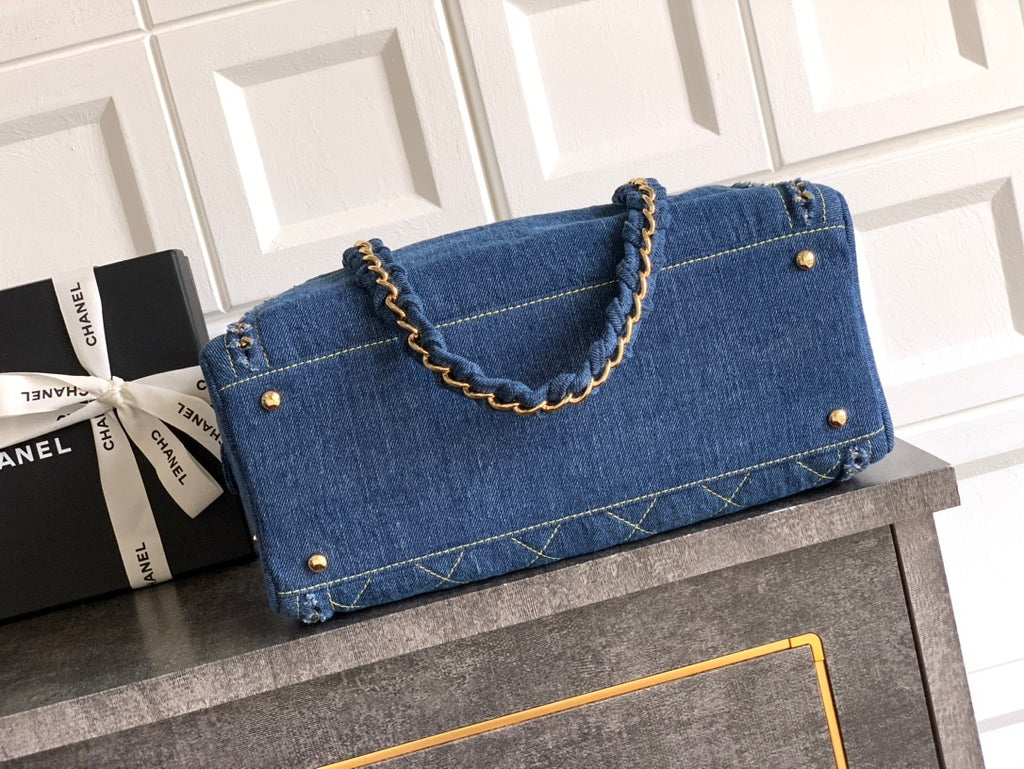 Blue denim handbag with gold accents on a gray surface next to a Chanel gift box.
