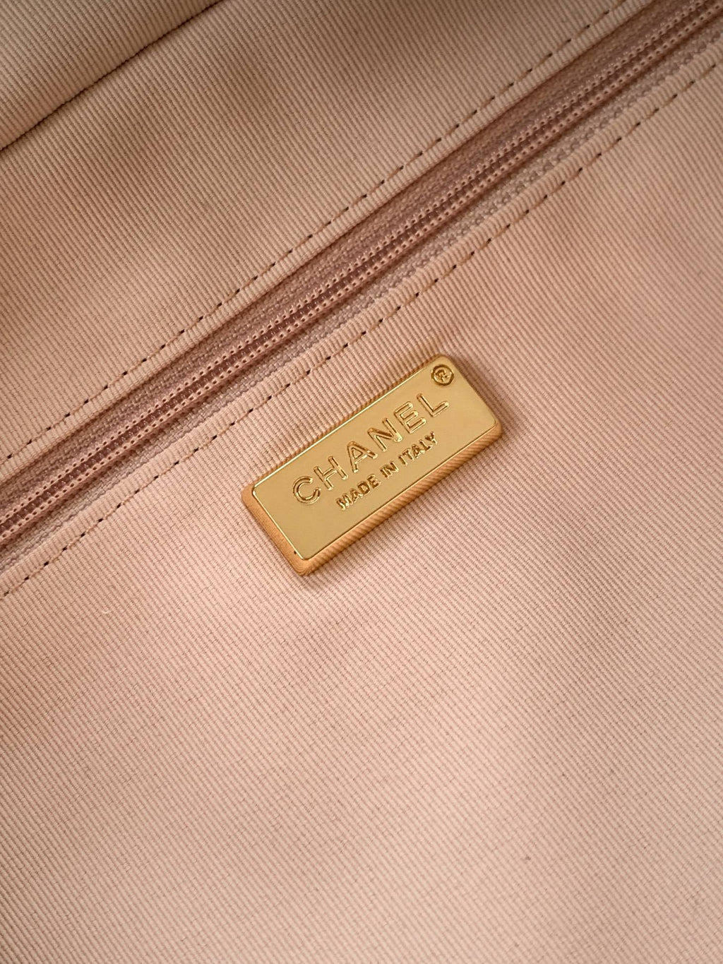 Close-up of a Chanel metal logo plaque on a light beige canvas bag, next to a matching zipper.