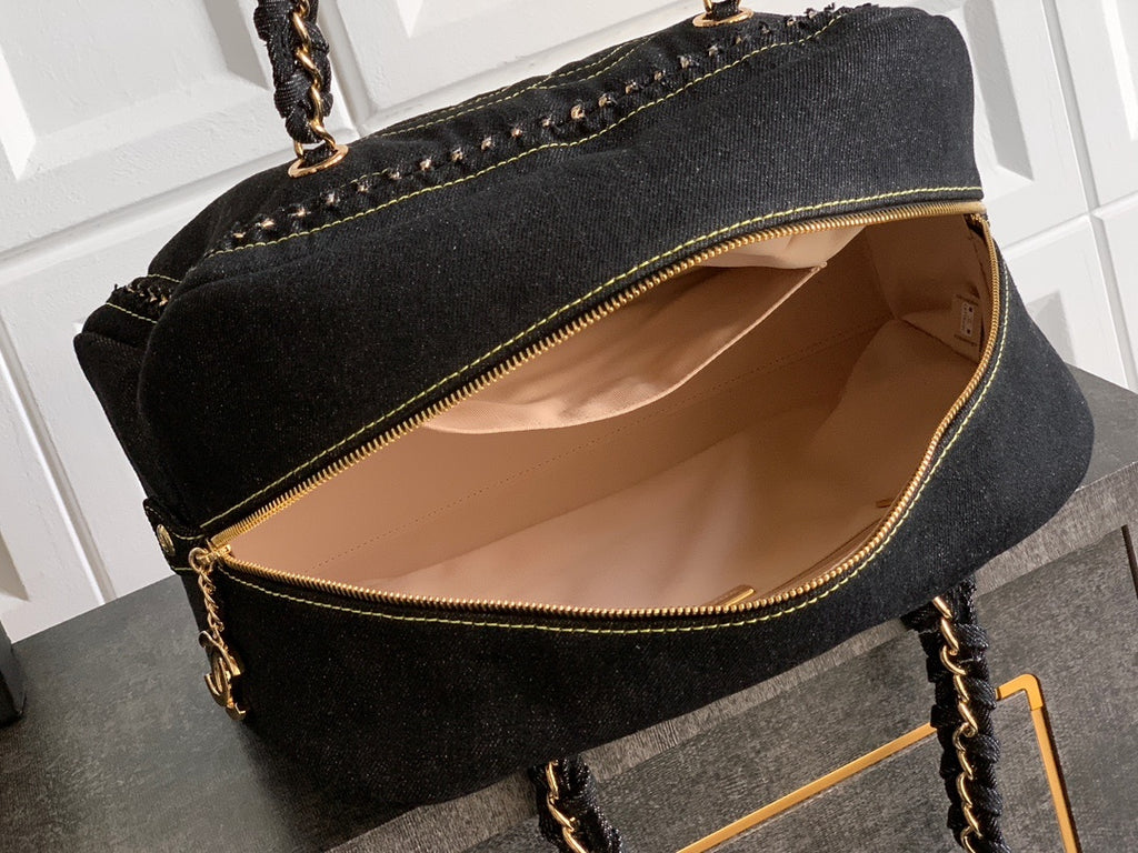 A black denim tote bag with gold-tone hardware, shown open to reveal a beige interior lining, resting on a dark surface against a white paneled background.