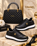 A pair of black Chanel low-top sneakers with quilted leather panels, gold-tone CC logos, and white soles, displayed alongside a black Chanel handbag on a beige marble-style set with gold accents.