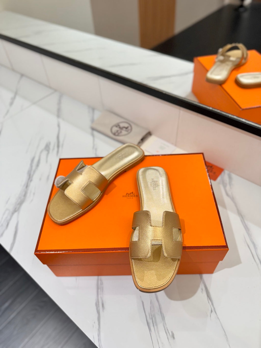 Gold sandals on an orange Hermès box with a marble surface.