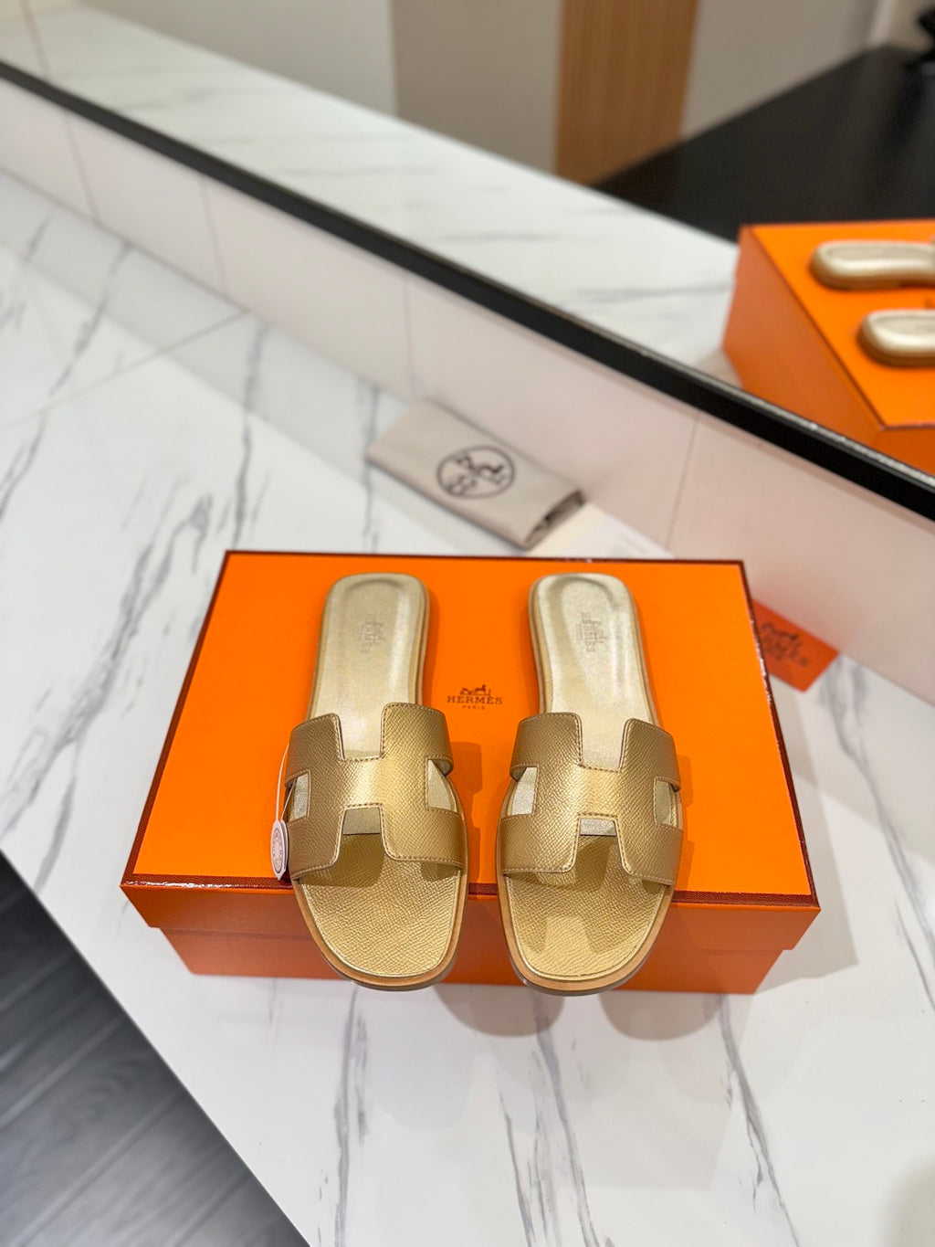 Gold sandals on an orange Hermès box with a marble floor background