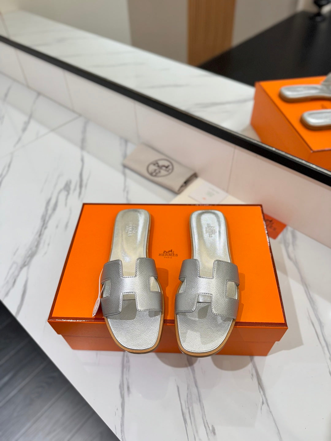 Silver sandals on an orange Hermès box with a mirror reflection.