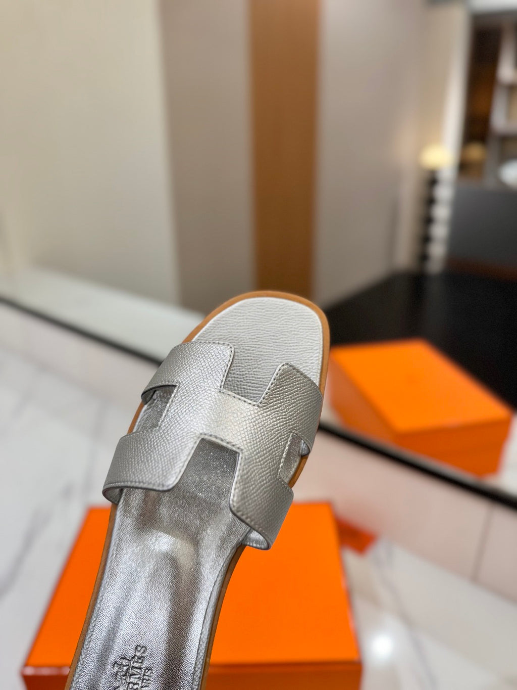 Silver sandal with a logo on an orange box in a blurred indoor setting
