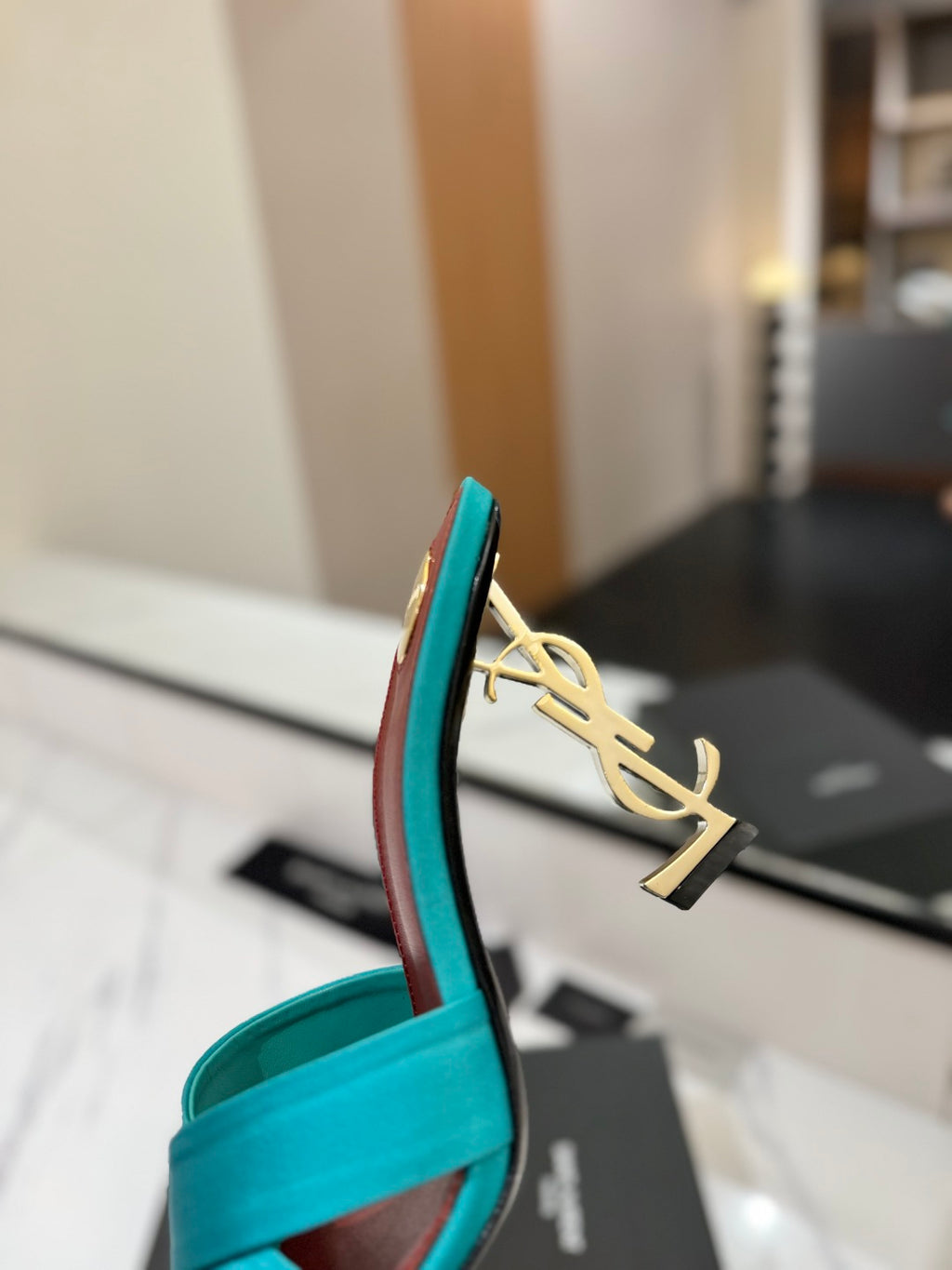 A close-up of a turquoise blue flat sandal with a gold-tone YSL monogram charm on the heel, resting on a black shoebox in a blurred indoor setting.