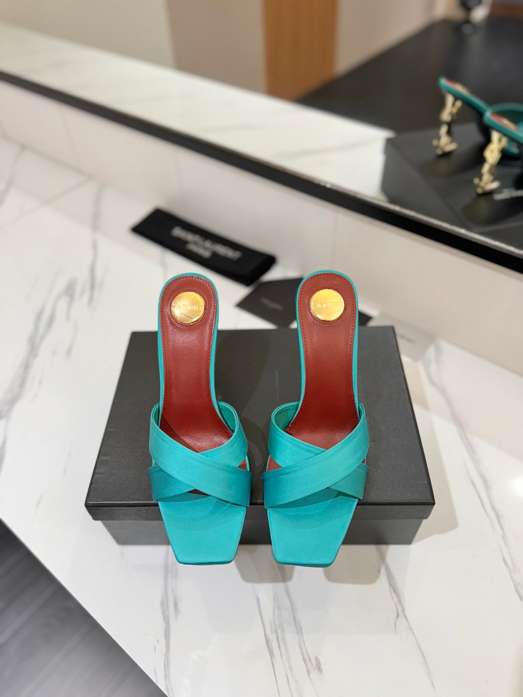A pair of turquoise slingback mule heels with crisscross straps and a block heel, displayed on a black shoebox on a white marble countertop. The insole is reddish-brown with a gold circular accent.