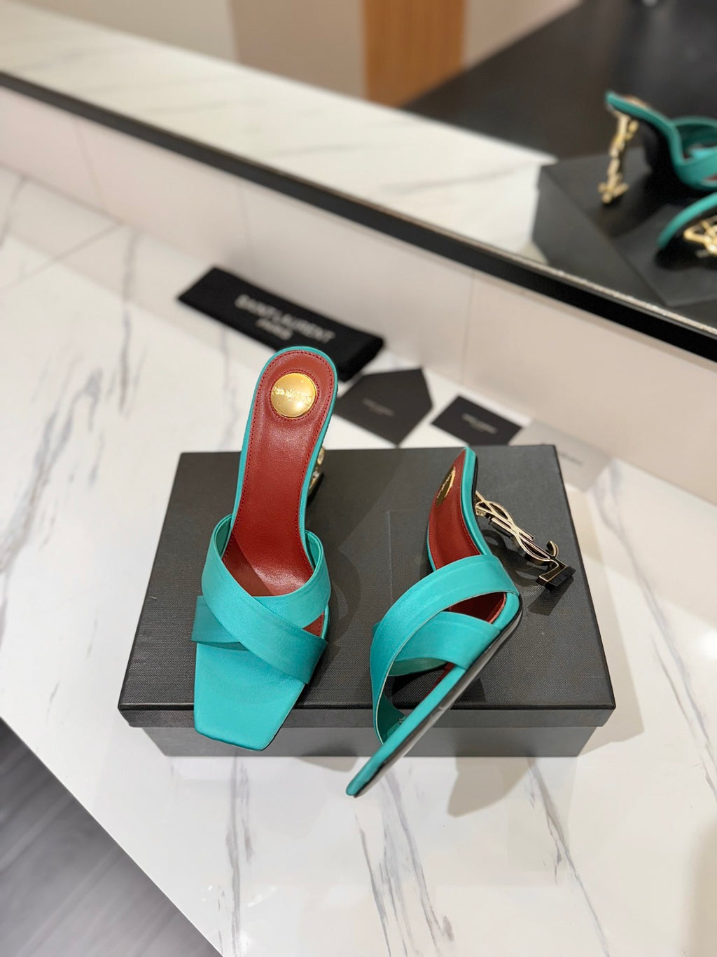 A pair of turquoise slingback mule heels with a red insole and gold heel cap, displayed on a black box on a white marble countertop.