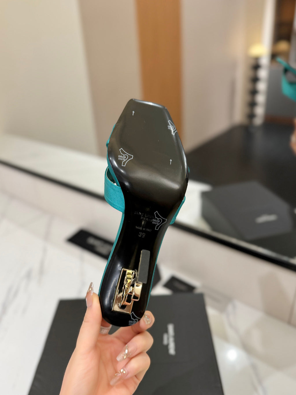 A hand holds a turquoise and black high-heeled shoe showing the sole, with a blurred indoor background and a black shoebox below.