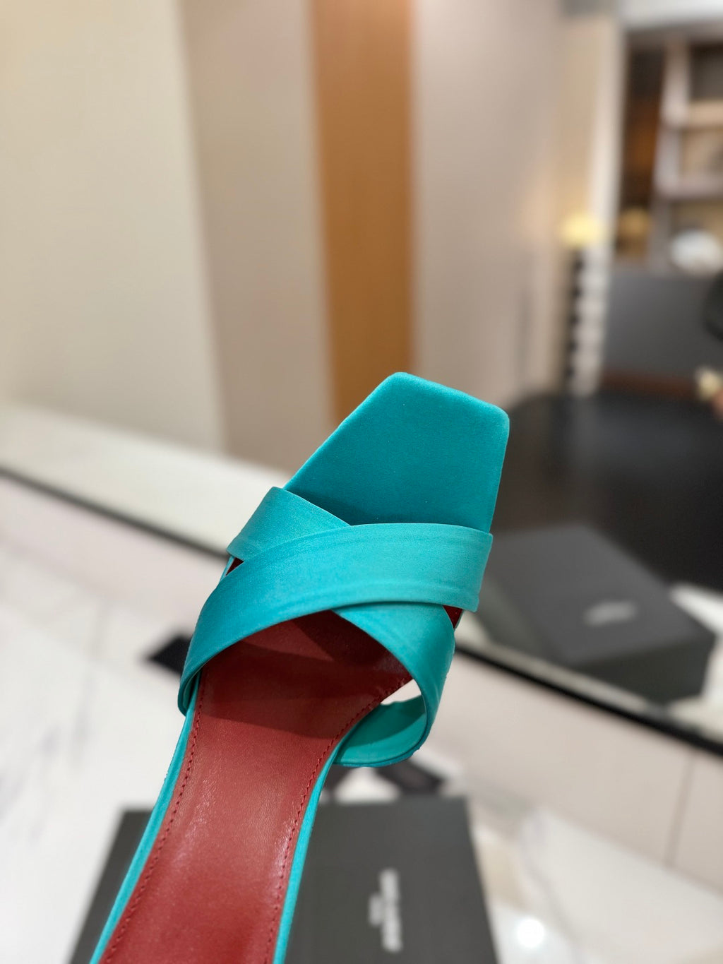 A close-up of a turquoise blue heeled sandal with a square toe and crisscrossing straps, resting on a white surface with a blurred indoor background.