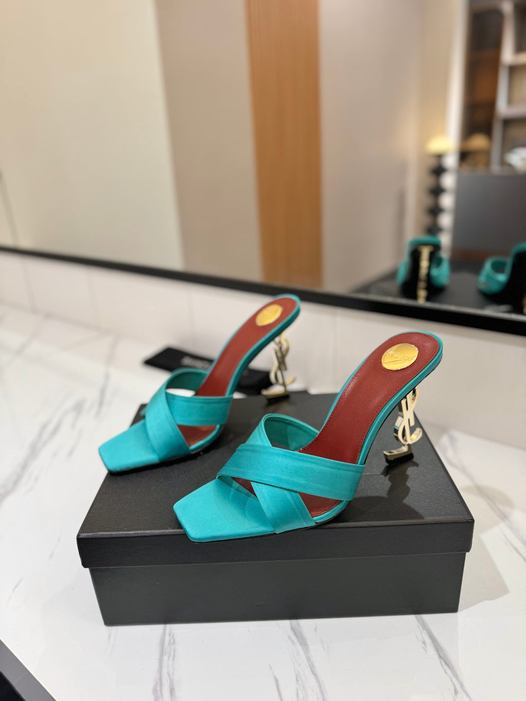 A pair of turquoise satin mule heels with metallic gold block heels and a gold circular emblem on the insole, displayed on a black shoe box on a marble countertop.