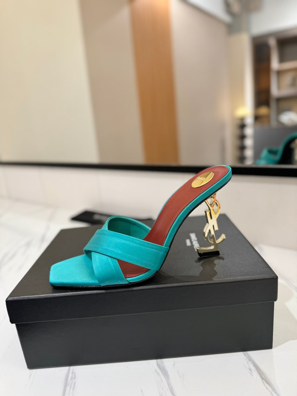 A turquoise satin slide sandal with a gold-tone YSL monogram heel, resting on a black Saint Laurent shoebox.
