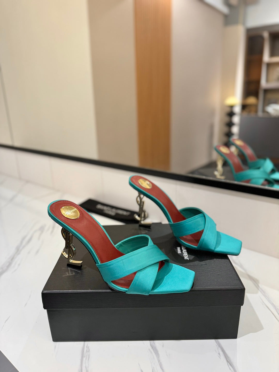 A pair of turquoise satin mule sandals with gold-tone high heels and red insoles, displayed on a black shoe box on a marble surface with a mirror in the background.