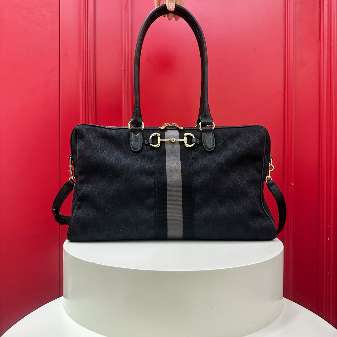 A black Gucci monogram canvas handbag with a central gray stripe and gold-tone horsebit hardware, displayed on a white cylindrical pedestal against a red paneled background.