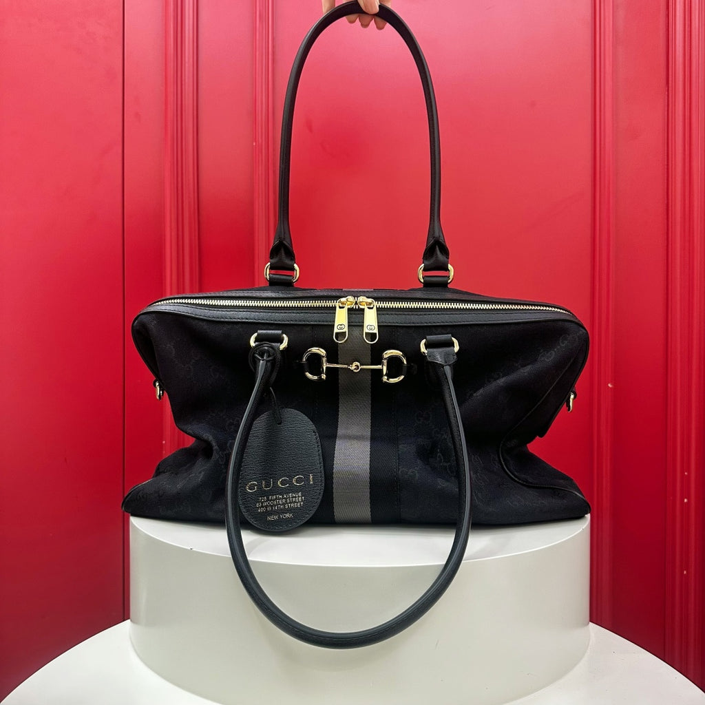A black Gucci horsebit shoulder bag with gold-tone hardware, displayed on a white pedestal against a bright red paneled background.