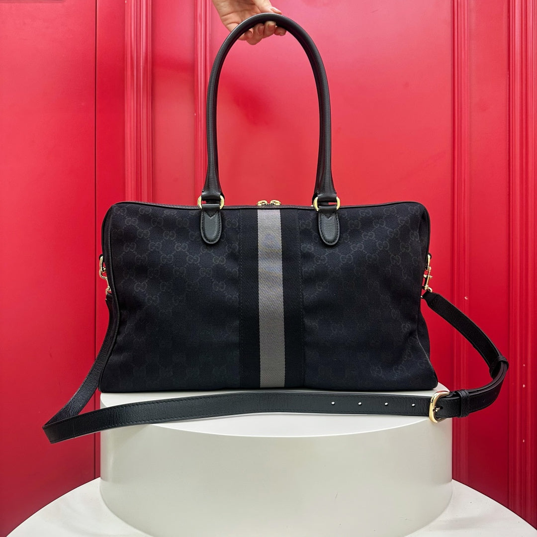 A black Gucci monogram satchel handbag with a central gray stripe, displayed on a white pedestal against a red paneled background. A hand is visible holding the top handle.