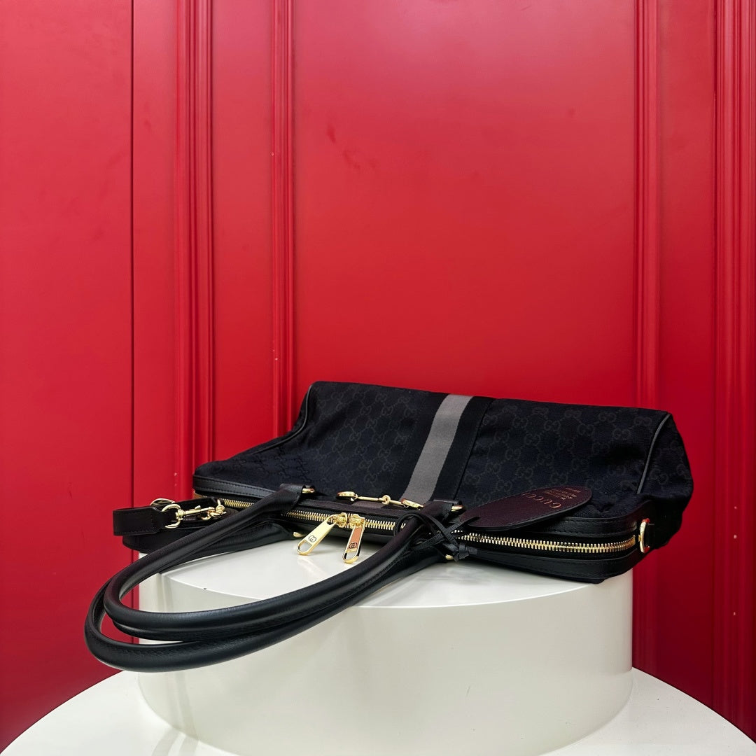 A black Gucci monogram clutch bag with gold-tone hardware and a dark brown leather logo tab, displayed on a white cylindrical pedestal against a vibrant red paneled background.