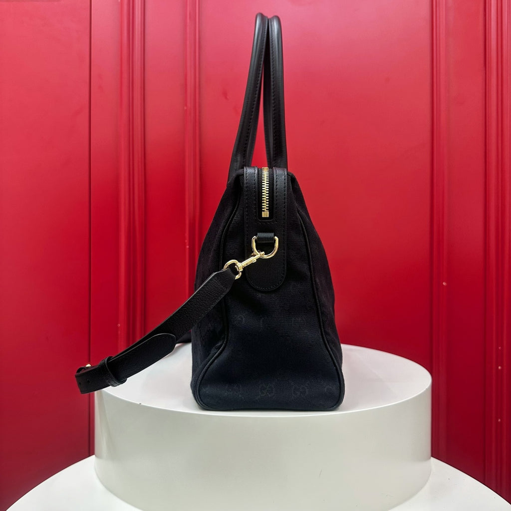 A black leather handbag with gold-tone hardware, displayed on a white cylindrical pedestal against a vibrant red paneled background.