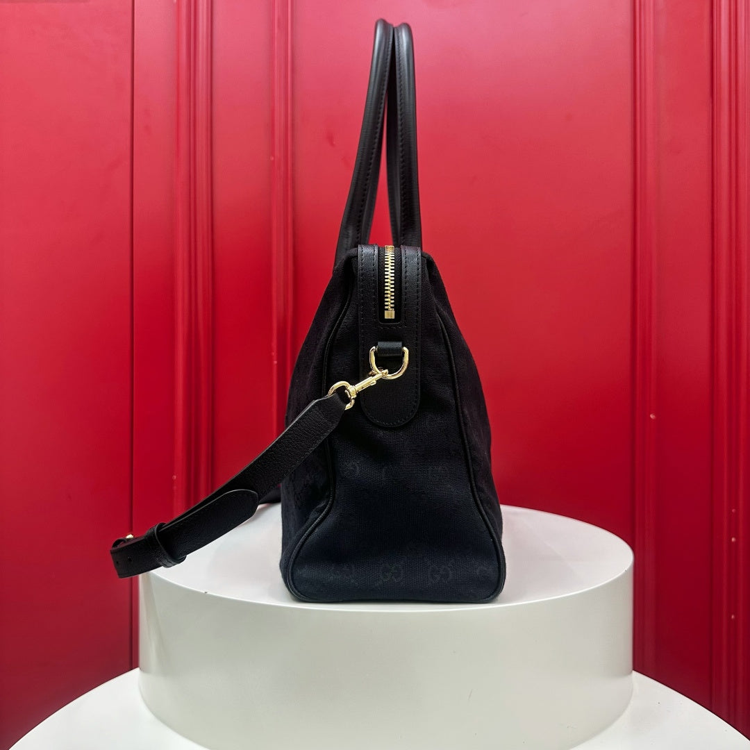 A black leather handbag with gold-tone hardware, displayed on a white cylindrical pedestal against a vibrant red paneled background.