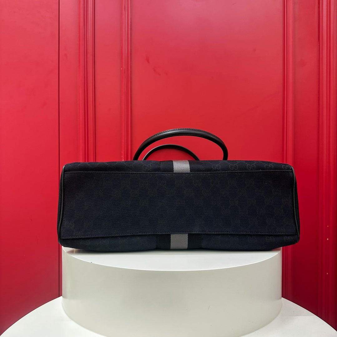 A black monogram canvas duffel bag with a dark gray leather handle and a gray vertical stripe, displayed on a white cylindrical pedestal against a bright red paneled background.