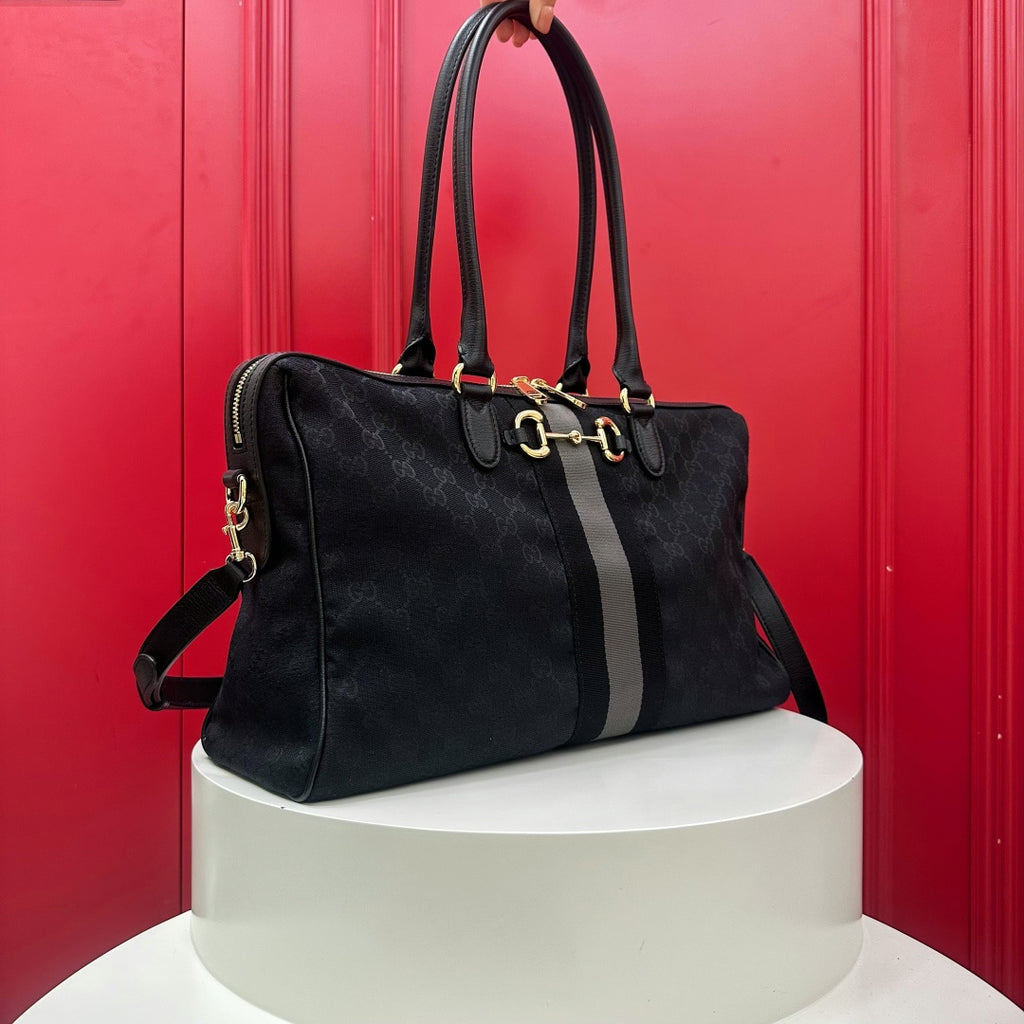 A black monogram canvas tote bag with a gray vertical stripe and gold horsebit hardware, held by a hand and displayed on a white cylindrical pedestal against a red paneled background.