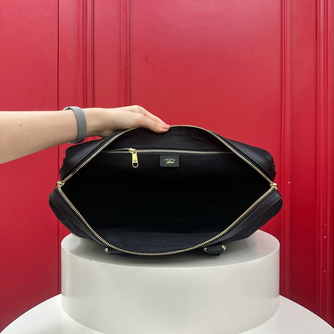 A black nylon laptop bag with gold zippers, shown open with a hand holding the top edge to display the interior. The bag is resting on a white cylindrical pedestal in front of a red paneled background.