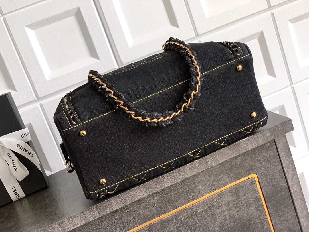 A black denim handbag with gold-tone hardware and a braided gold-and-black handle, displayed on a gray cabinet against a white paneled wall.