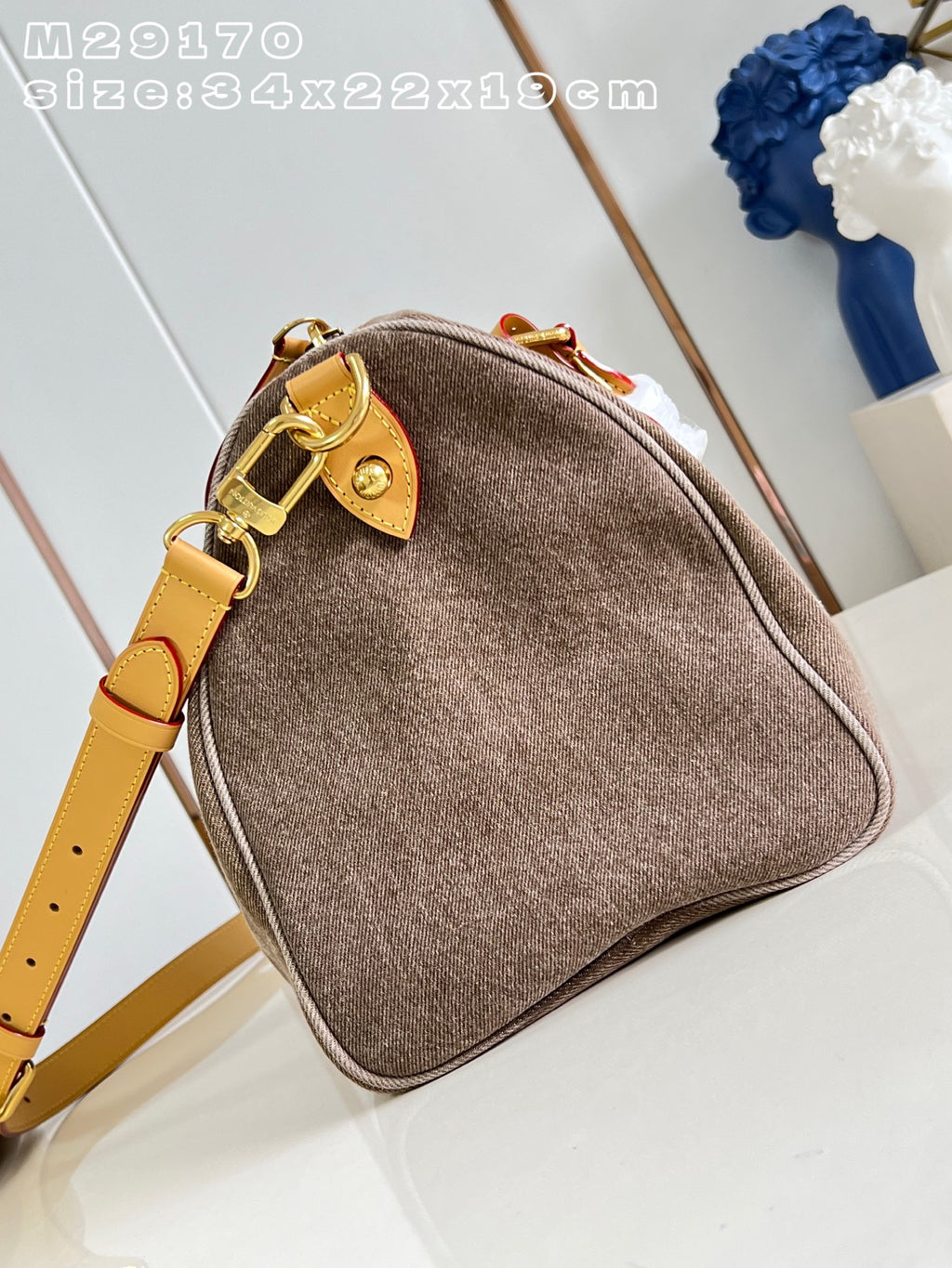 A brown felt shoulder bag with a tan leather strap and gold-tone hardware, shown on a light surface with a decorative blue flower in the background. White text in the top left reads 'M29170 size:34x22x19cm'.