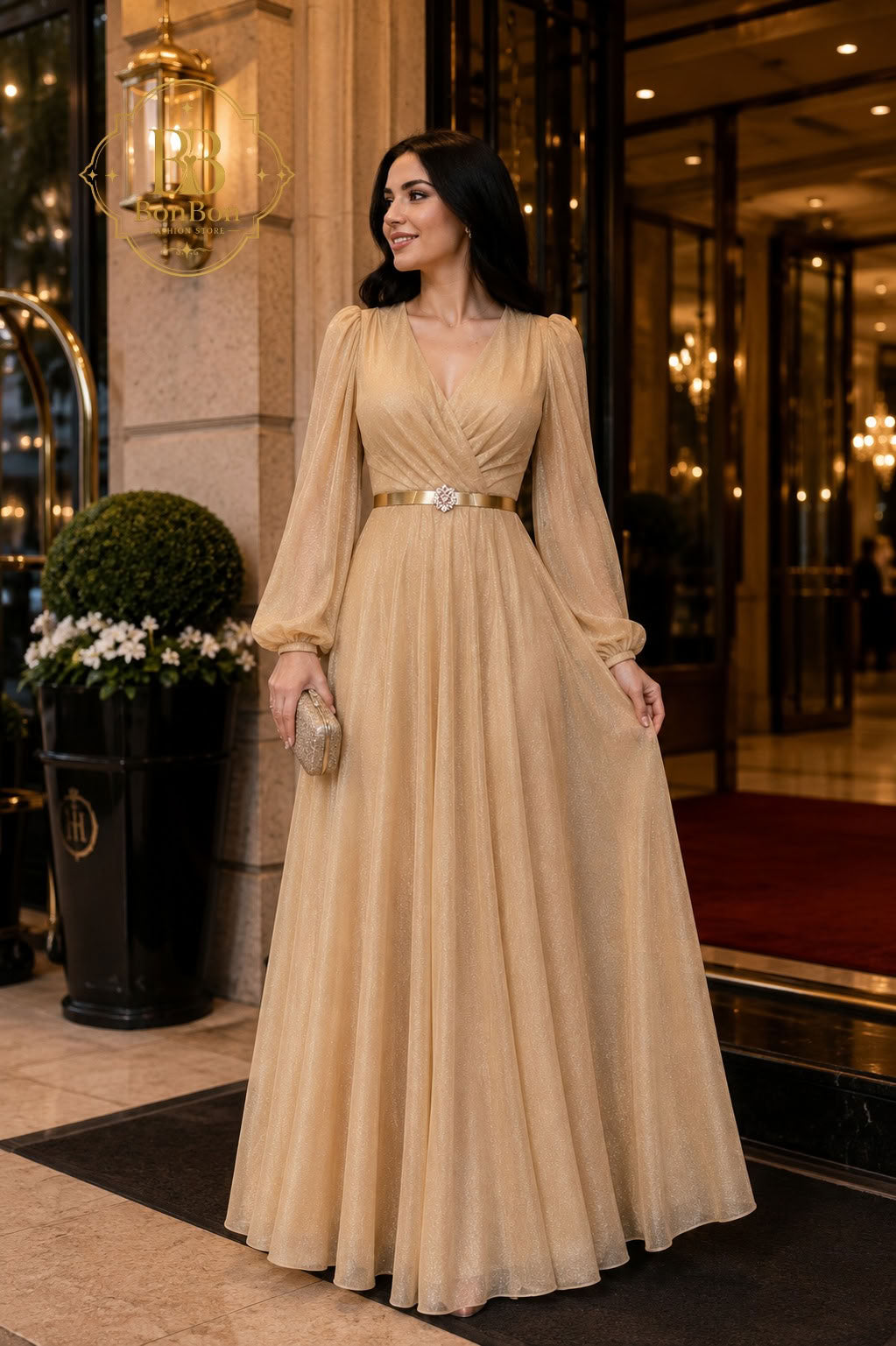 A woman wearing a floor-length champagne beige gown with long sheer sleeves and a metallic gold belt, standing in front of a luxury hotel entrance.