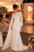 A woman wearing a white off-the-shoulder mermaid-style wedding gown with a long sheer cape and lace sleeves, standing in an outdoor event space at sunset with tables, flowers, and warm string lights in the background.