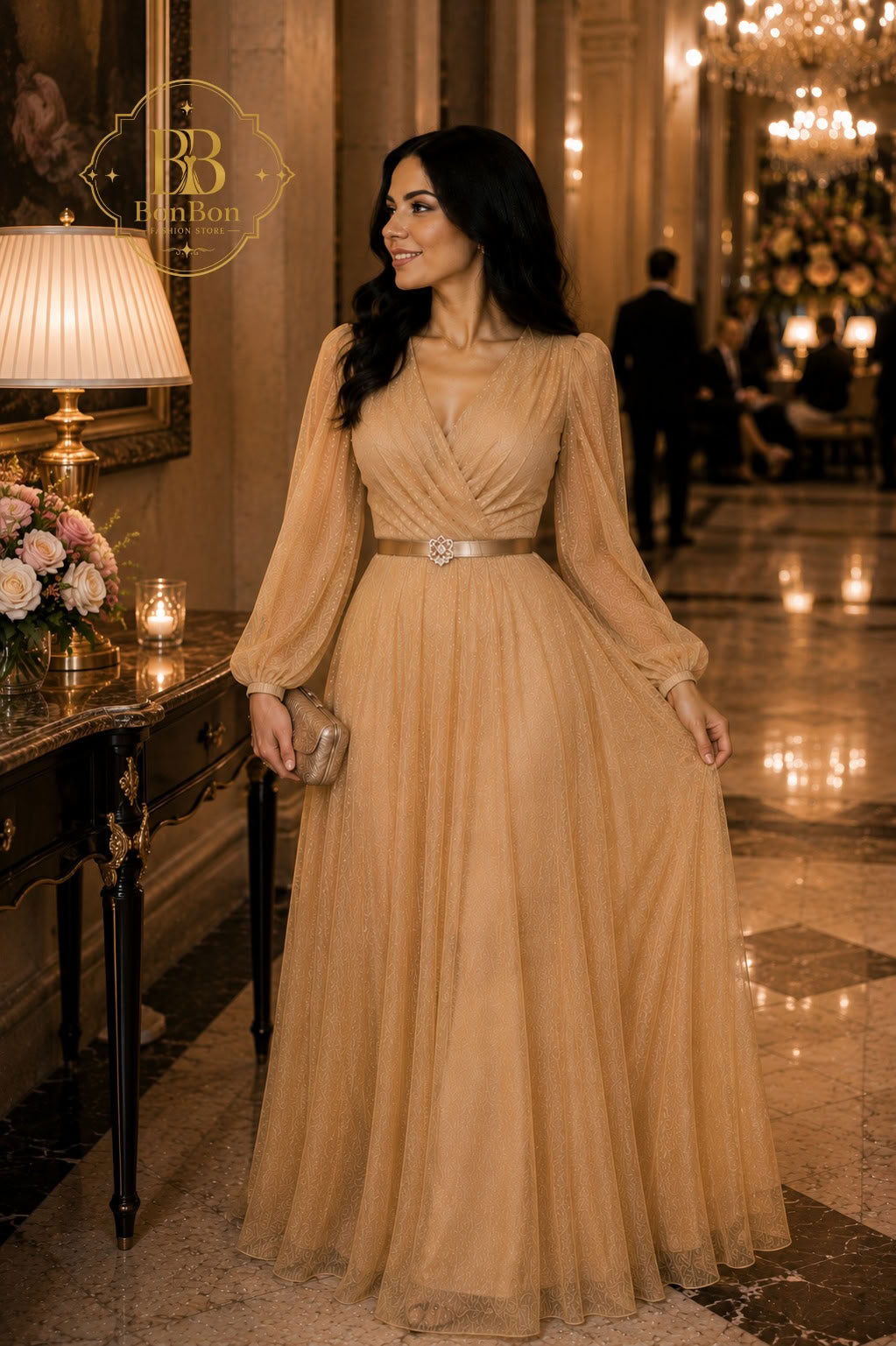 A woman wearing a floor-length champagne beige gown with long sheer sleeves and a silver waistband, standing in an elegant indoor hallway with warm lighting and a chandelier in the background.