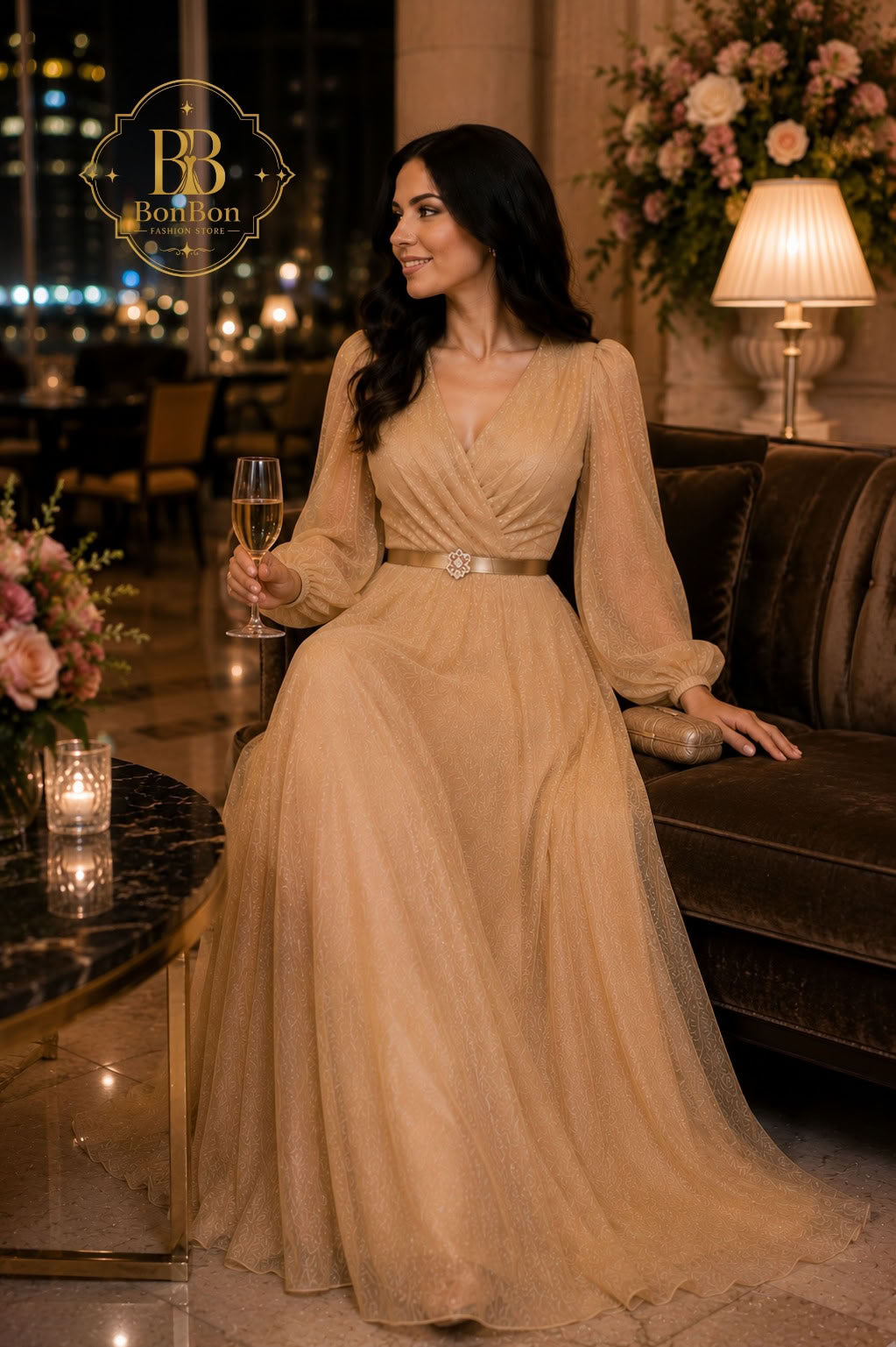 A woman wearing a floor-length champagne beige gown with long sheer sleeves and a silver belt sits on a dark velvet sofa in a luxurious room, holding a glass of champagne. A 'BB Fashion' logo is visible in the top left corner.