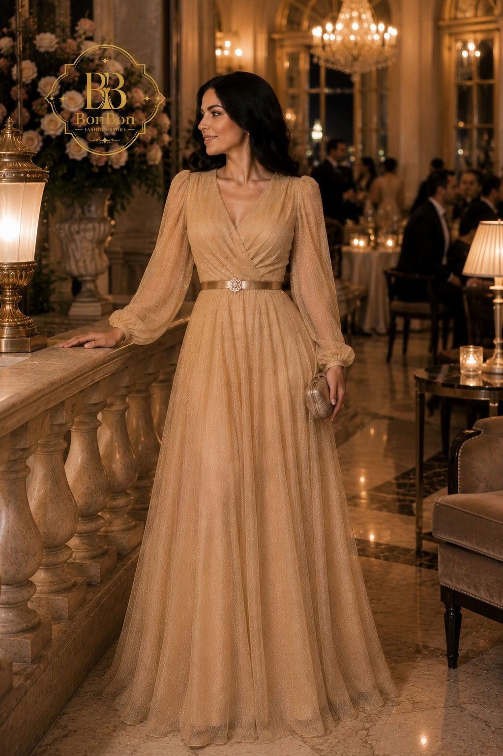 A woman wearing a floor-length champagne beige gown with long sheer sleeves and a thin metallic belt stands in an elegant indoor event space with chandeliers and guests in the background.