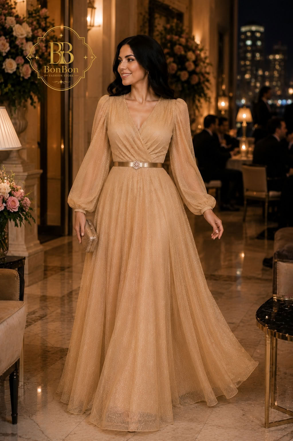 A woman wearing a floor-length champagne beige gown with long sheer puff sleeves and a thin metallic belt, standing in a warmly lit indoor event space. The image includes a visible 'BB BonBon' logo watermark in the upper left.