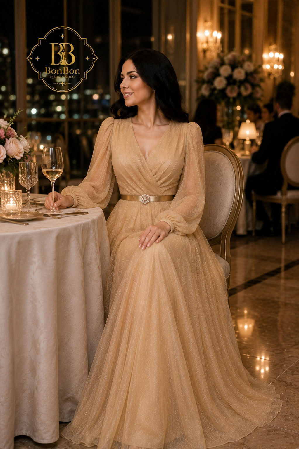 A woman wearing a champagne beige long-sleeve tulle evening gown with a silver belt, seated at a table in an elegant restaurant setting. A 'BonBon' brand logo is visible in the top left corner.