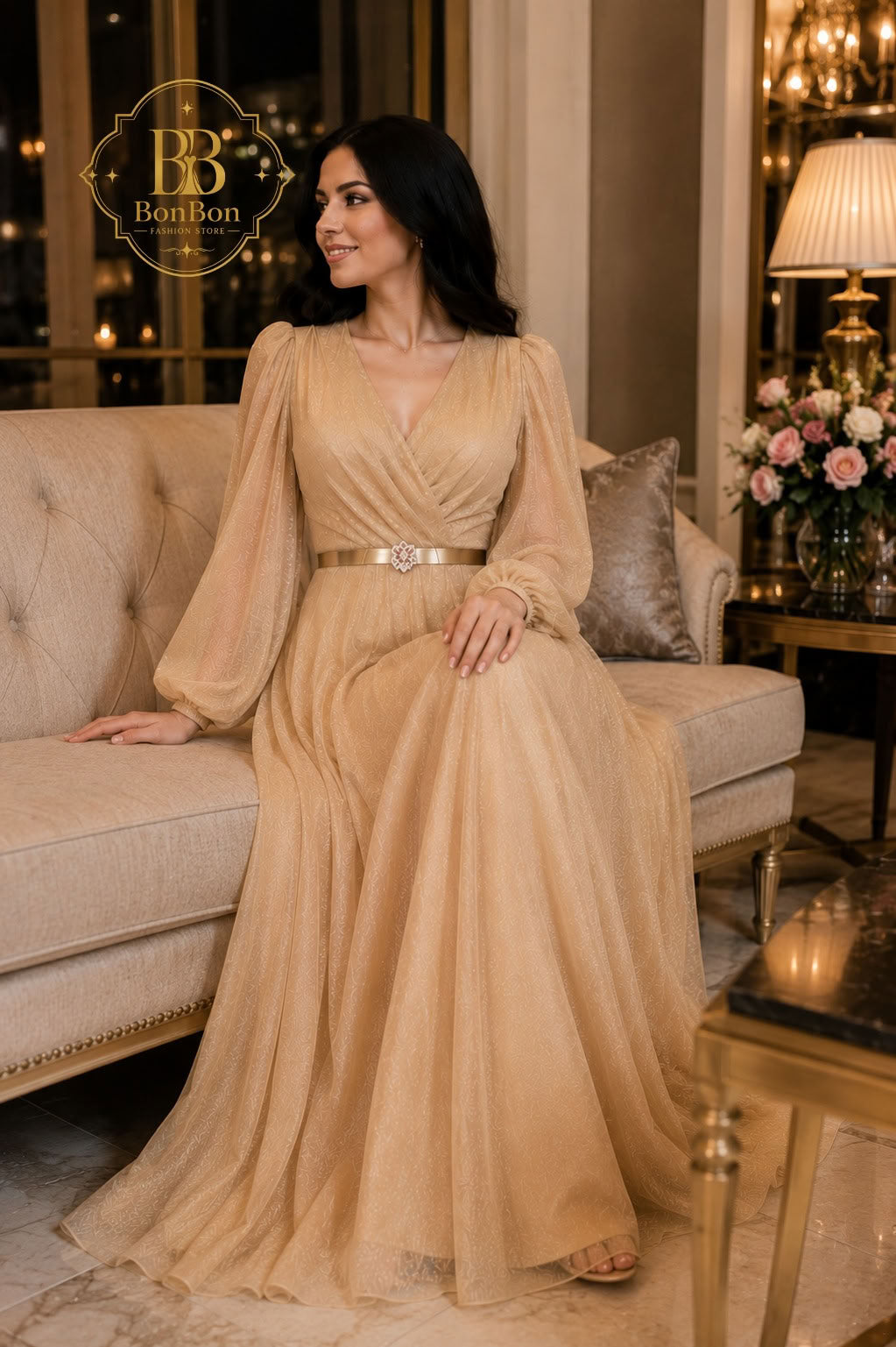 A woman wearing a floor-length champagne beige gown with long sheer sleeves and a metallic belt, seated on a beige sofa in an elegant indoor setting. The BonBon brand logo is visible in the top left corner.