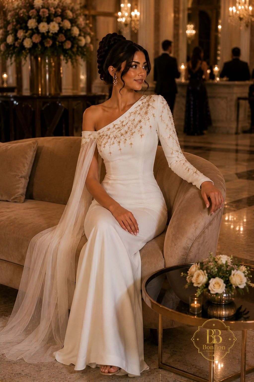 A woman wearing an elegant off-the-shoulder white wedding gown with long sheer lace sleeves and a flowing veil, seated on a beige sofa in a warmly lit indoor venue. A small round table with flowers sits beside her, and blurred guests are visible in the background.